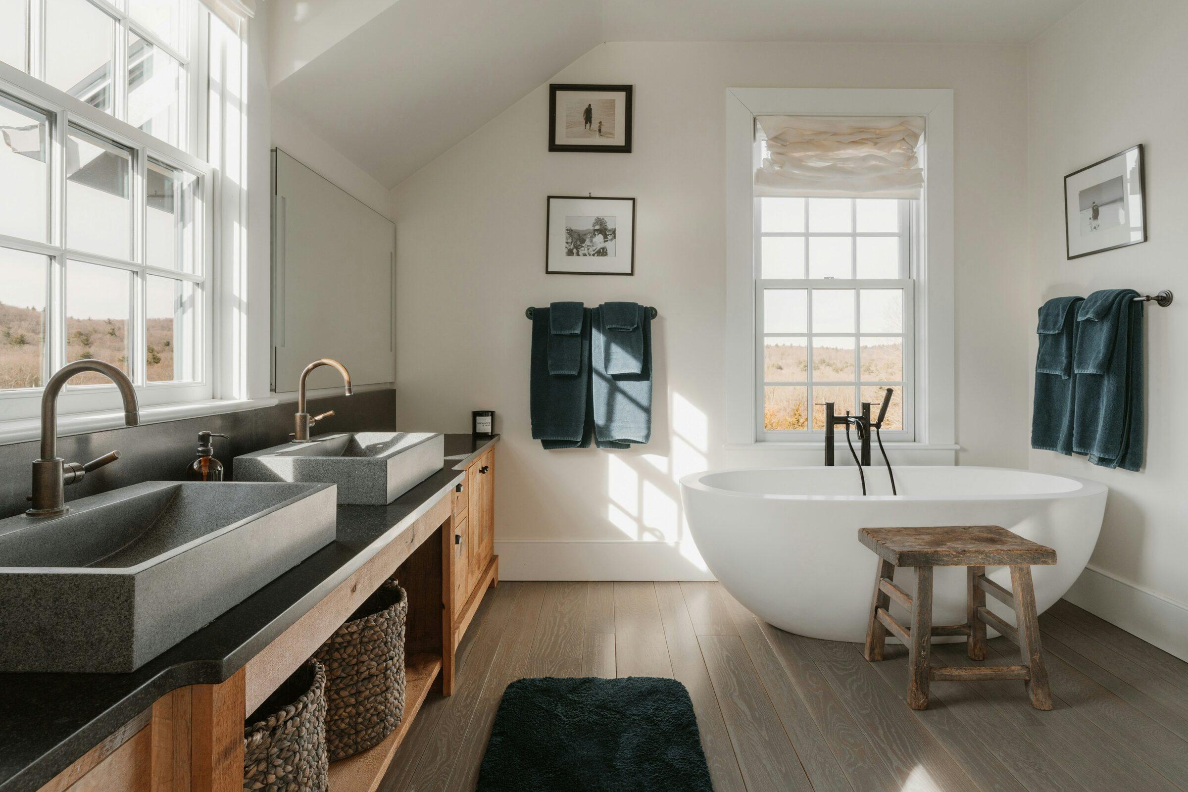 Bathroom with a white freestanding tub under a window, a wood vanity with two grey stone vessel sinks and bronze faucets, and grey wood-look floors