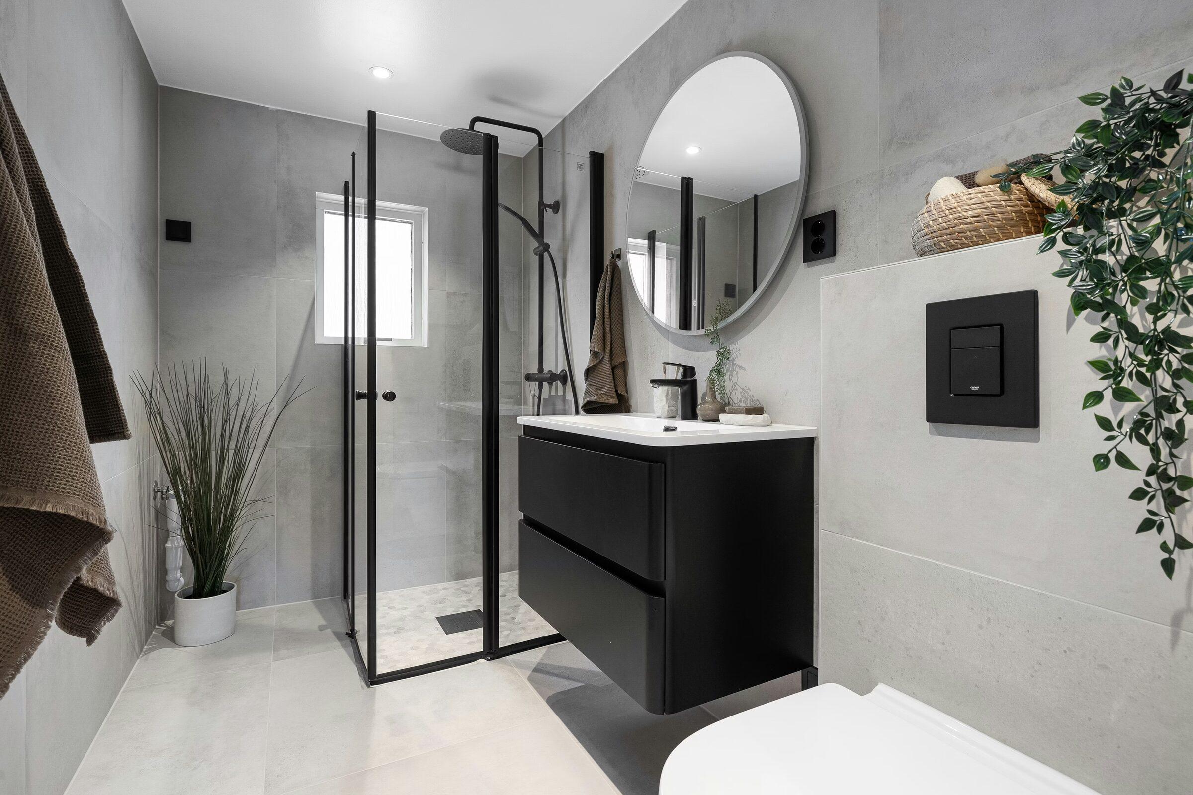 Bathroom with large grey tiles, a black-framed glass shower, a black floating vanity with a white basin, a round mirror and a wall-hung toilet