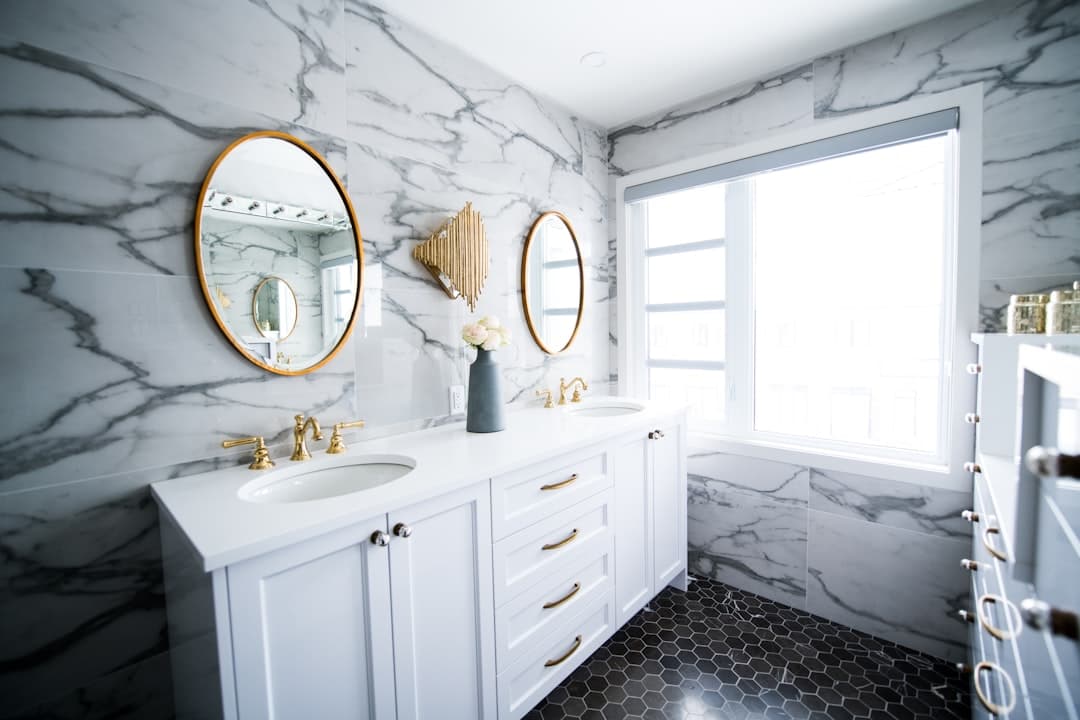 Bathroom with marble-look wall tile, a white double vanity with brass faucets and pulls, two round gold-framed mirrors and black hexagon floor tile