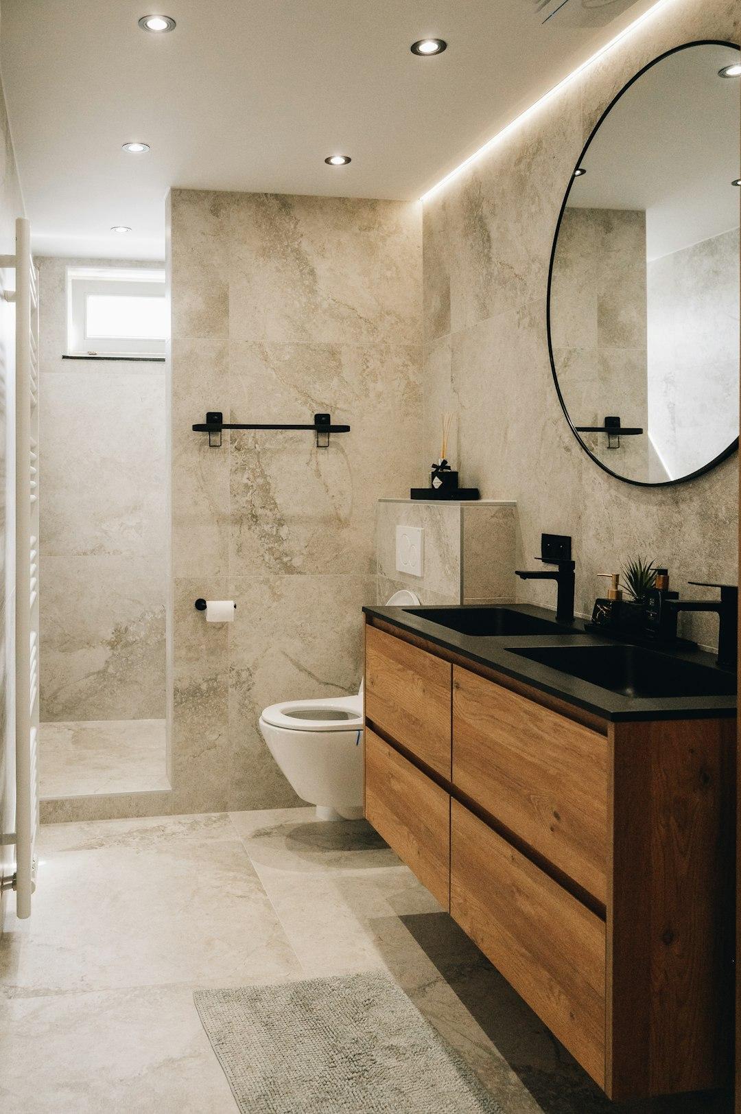 Bathroom with beige stone-look tile, a wood floating vanity with a black double basin and black faucets, a large round mirror and a wall-hung toilet