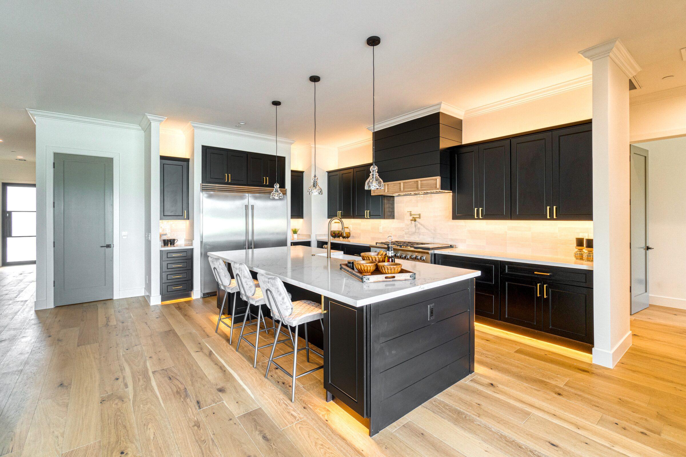 Open kitchen with black shaker cabinets, a white marble-look island with three bar stools, glass pendant lights and wide-plank wood floors
