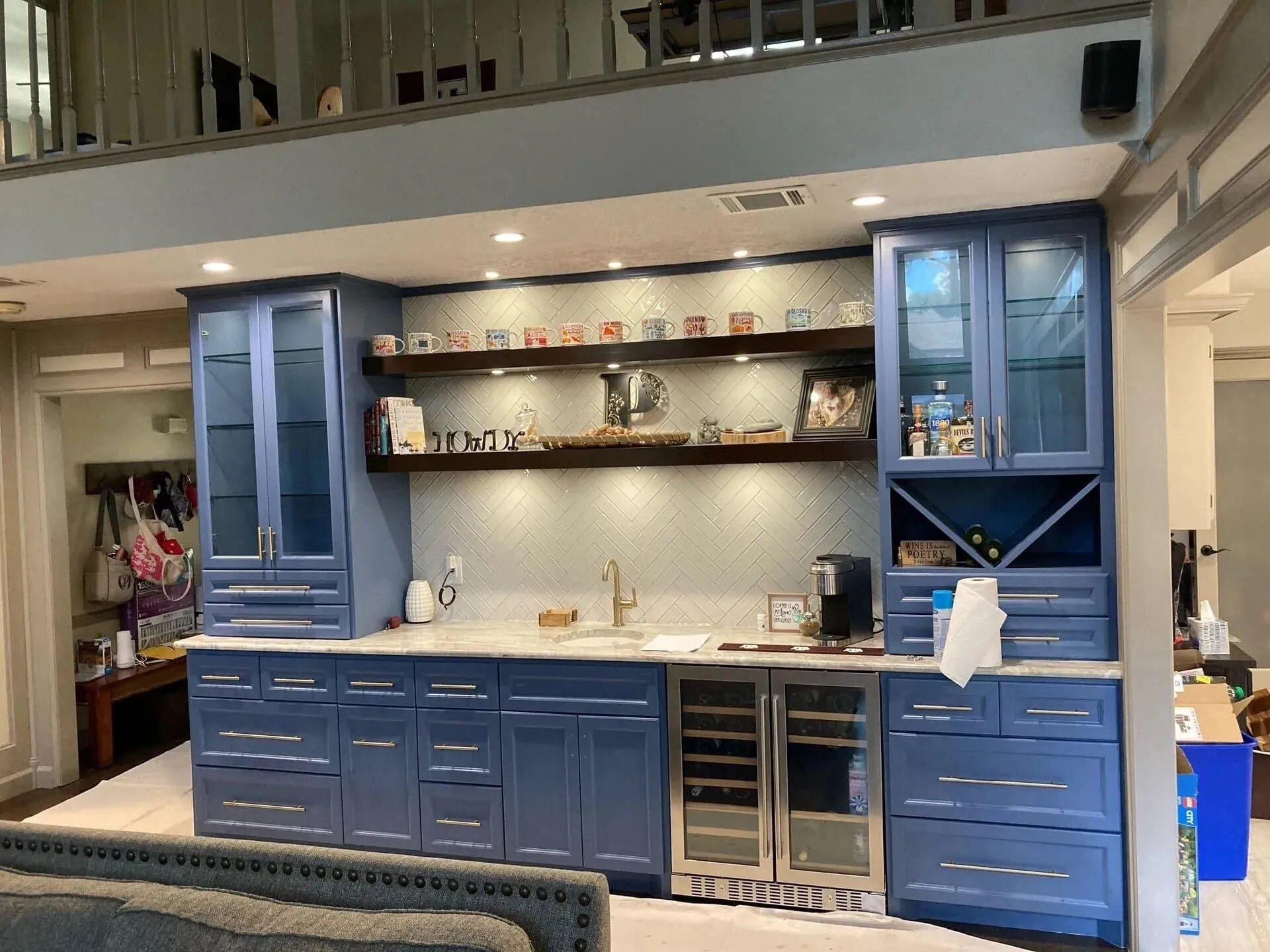Built-in bar wall with blue shaker cabinets, glass-front upper cabinets, dark floating shelves, a white herringbone tile backsplash, a small sink with a brass faucet and an under-counter wine fridge