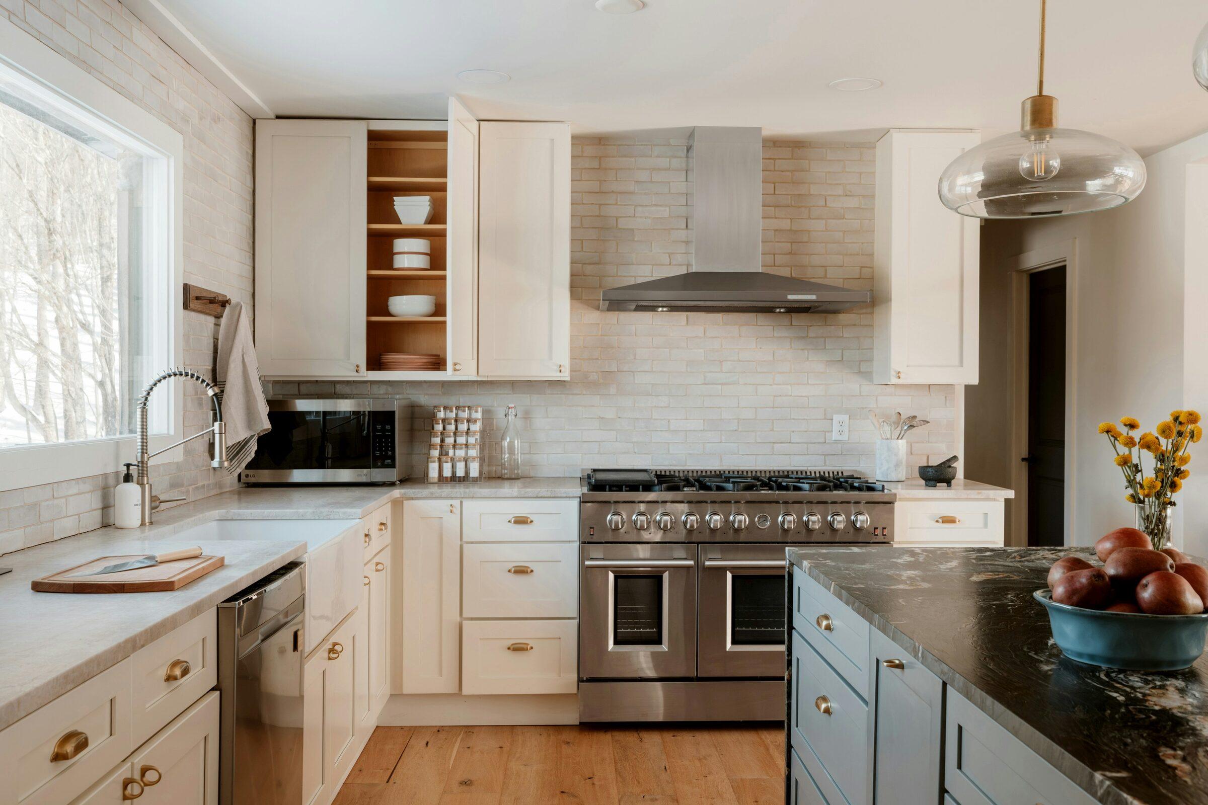 Kitchen with cream shaker cabinets and brass pulls, a stainless steel double-oven range and hood, a pale brick-pattern tile backsplash and a dark stone island