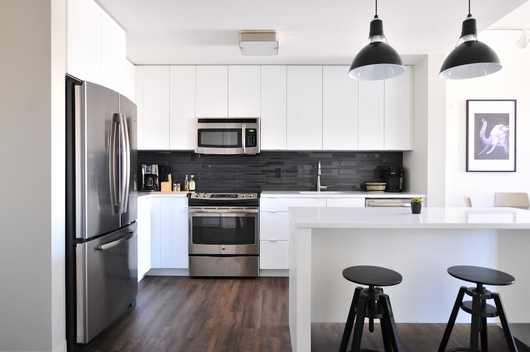 White kitchen with flat-panel cabinets, a dark tile backsplash, a stainless refrigerator and range, a white island with two black stools and two black pendant lamps