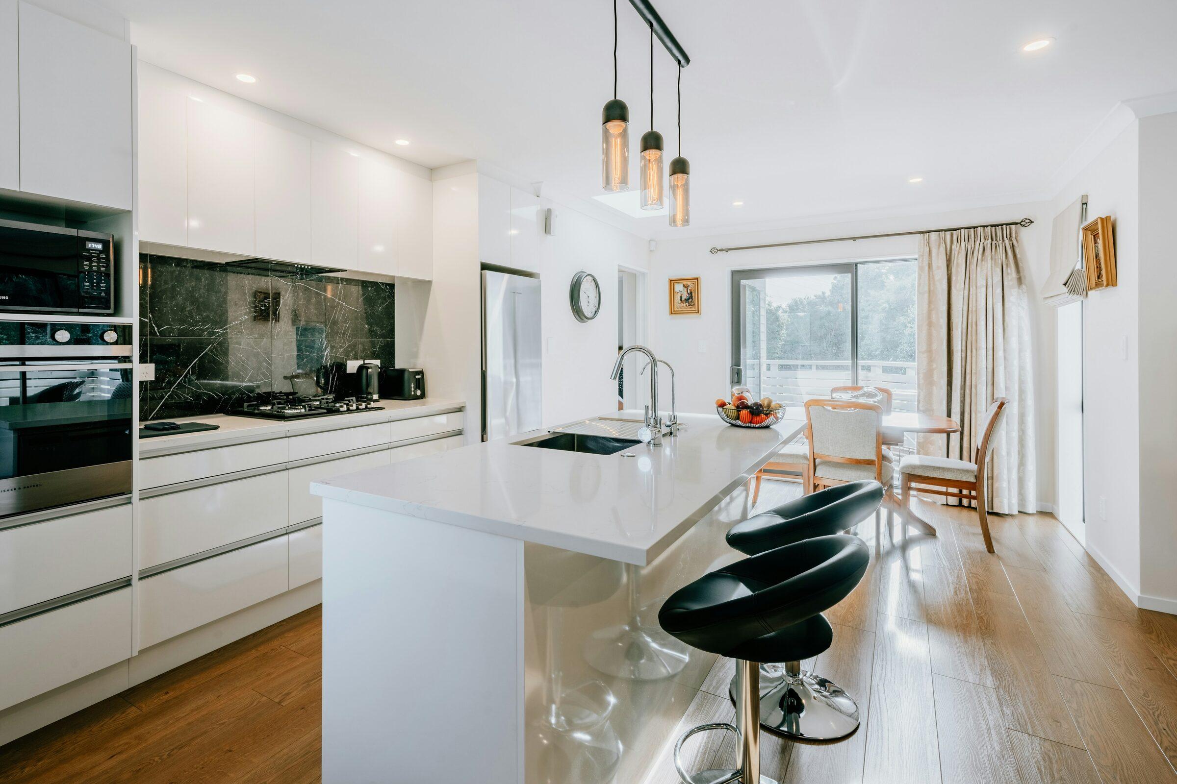 White gloss kitchen with a white island and sink, two black swivel stools, a black marble backsplash and wood floors opening onto a dining area