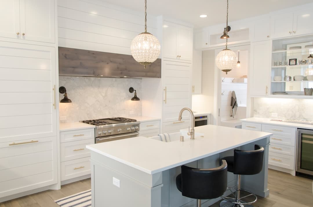 White kitchen with panelled cabinets, a marble backsplash behind a steel range, an island with a white countertop and farmhouse sink, and two crystal globe pendant lights